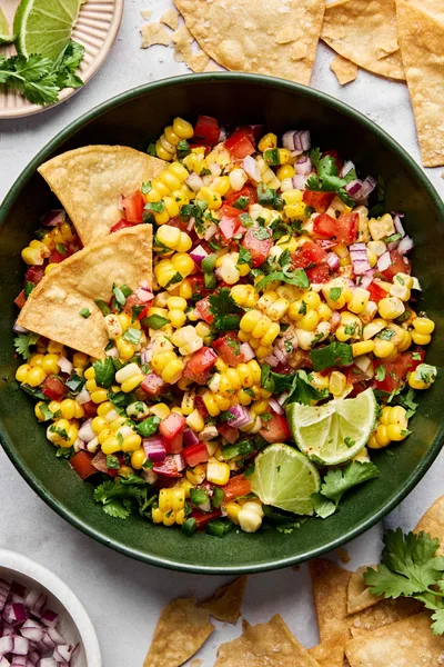 Bowl of fresh corn salsa with diced tomatoes, red onion, jalapeño, cilantro, and lime surrounded by crispy tortilla chips