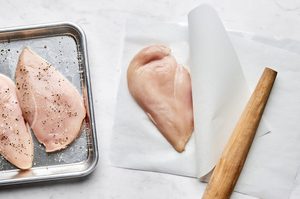 seasoned chicken breasts being pounded flat with a rolling pin between parchment paper to make a grilled chicken sandwich