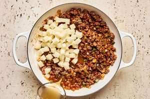 potatoes, chicken stock and spices being added to a skillet of cooked ground beef to make a heart one pot dinner