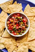 A vibrant tomato and olive salsa in a white bowl, featuring diced tomatoes, black olives, and chiles, served with chips