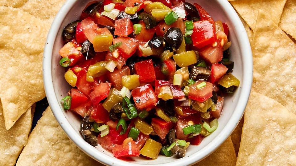 A vibrant tomato and olive salsa in a white bowl, featuring diced tomatoes, black olives, and chiles, served with chips