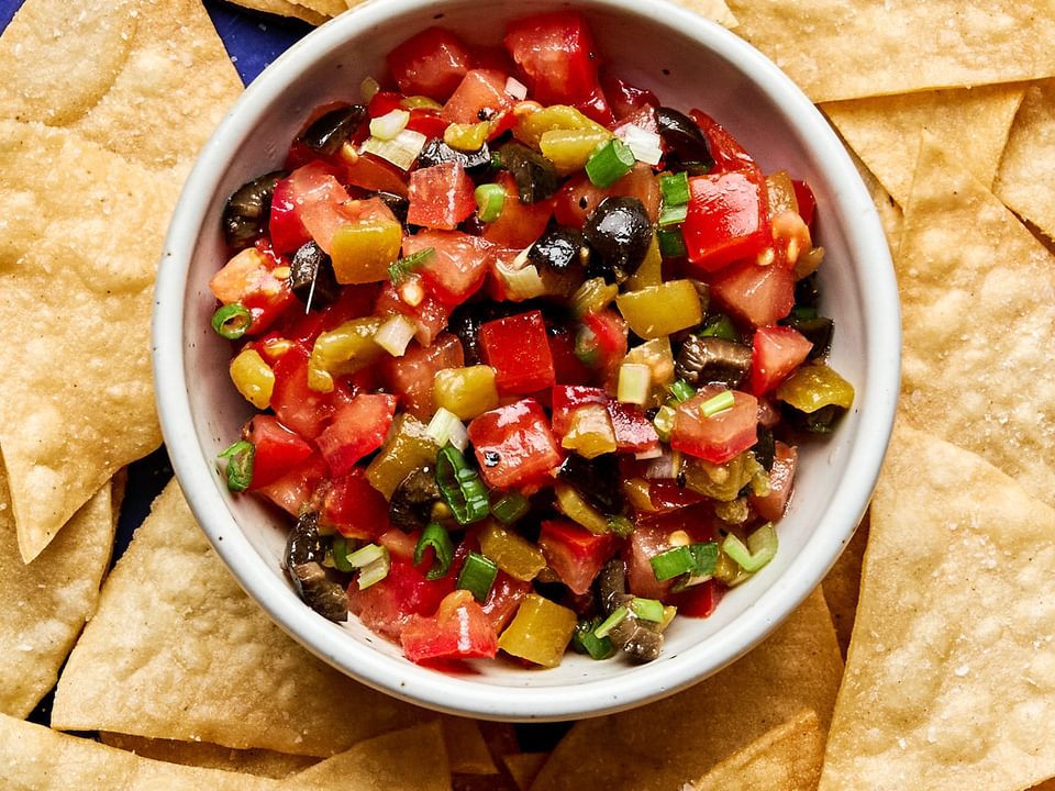 A vibrant tomato and olive salsa in a white bowl, featuring diced tomatoes, black olives, and chiles, served with chips