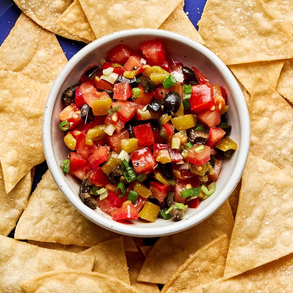 A vibrant tomato and olive salsa in a white bowl, featuring diced tomatoes, black olives, and chiles, served with chips