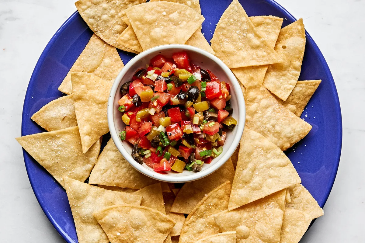 A vibrant tomato and olive salsa in a white bowl, featuring diced tomatoes, black olives, and chiles, served with chips