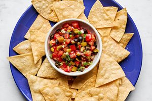 A vibrant tomato and olive salsa in a white bowl, featuring diced tomatoes, black olives, and chiles, served with chips