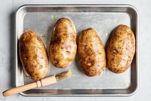 4 russet potatoes being brushed with olive oil on a rimmed baking sheet