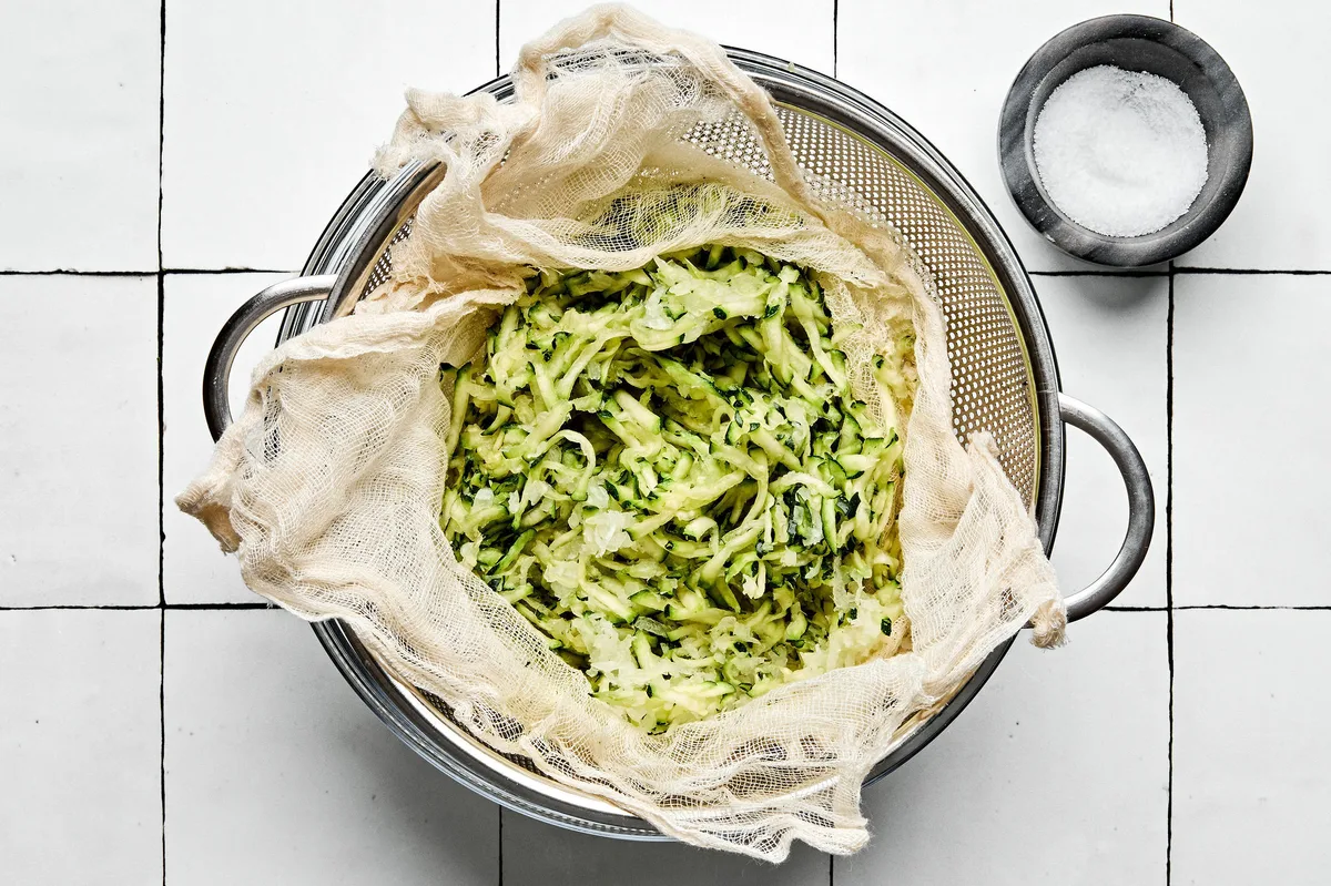 grated zucchini being strained through cheese cloth in a colander next to salt to help absorb moisture