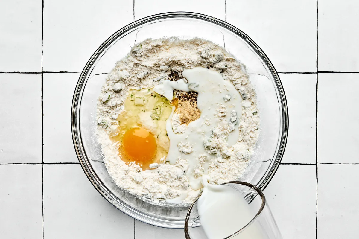 batter for zucchini fritters being made in a bowl with flour, baking powder, green onions, Parmesan, egg, milk and spices
