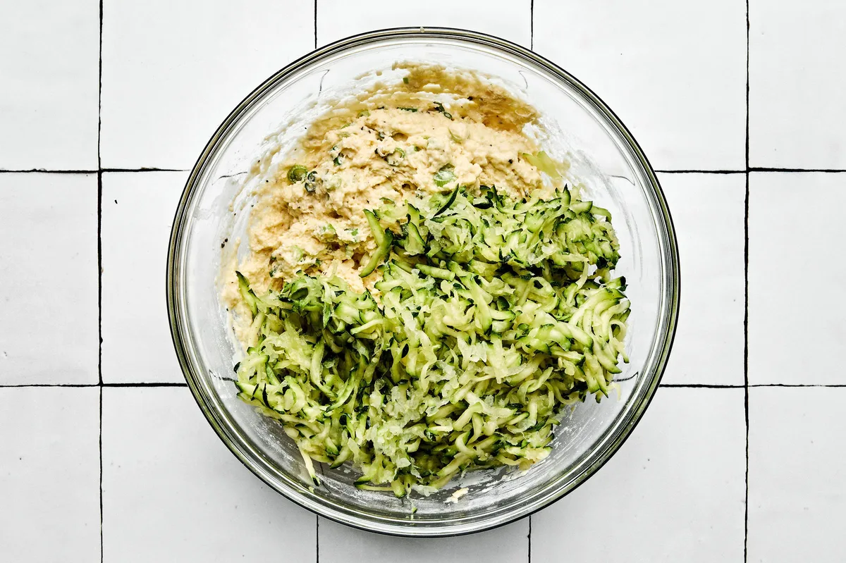 batter for zucchini fritters being made in a bowl with flour, green onions, Parmesan, egg, milk, spices and grated zucchini