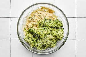 batter for zucchini fritters being made in a bowl with flour, green onions, Parmesan, egg, milk, spices and grated zucchini