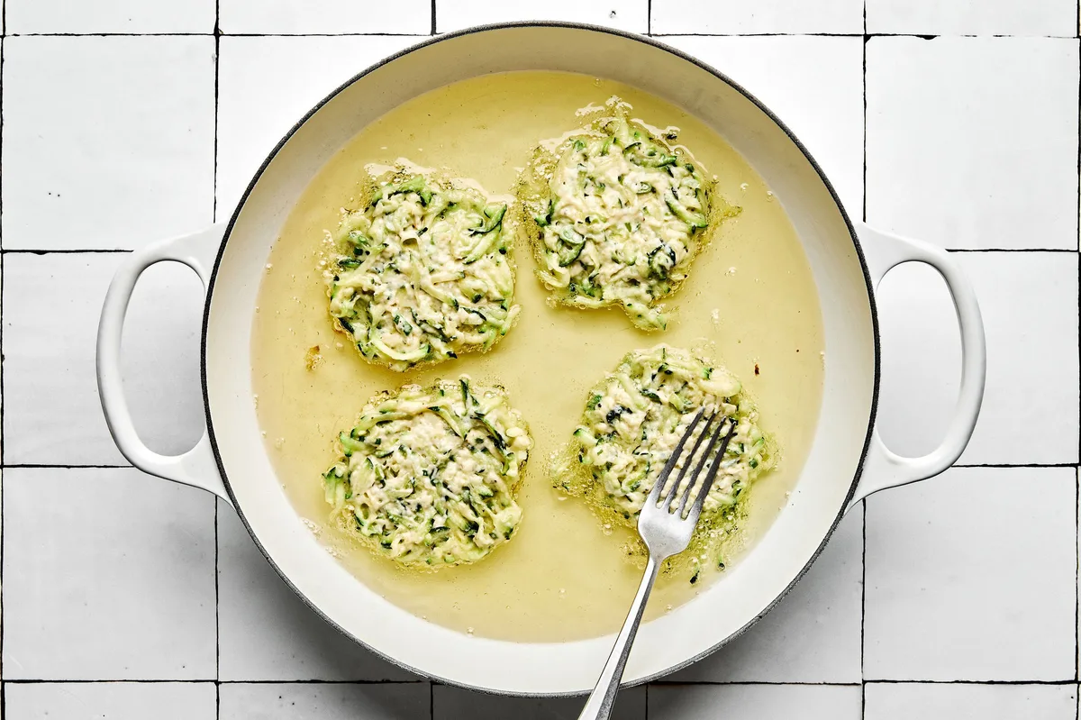 A skillet filled with sizzling oil, actively cooking several zucchini fritters until golden-brown with crispy edges