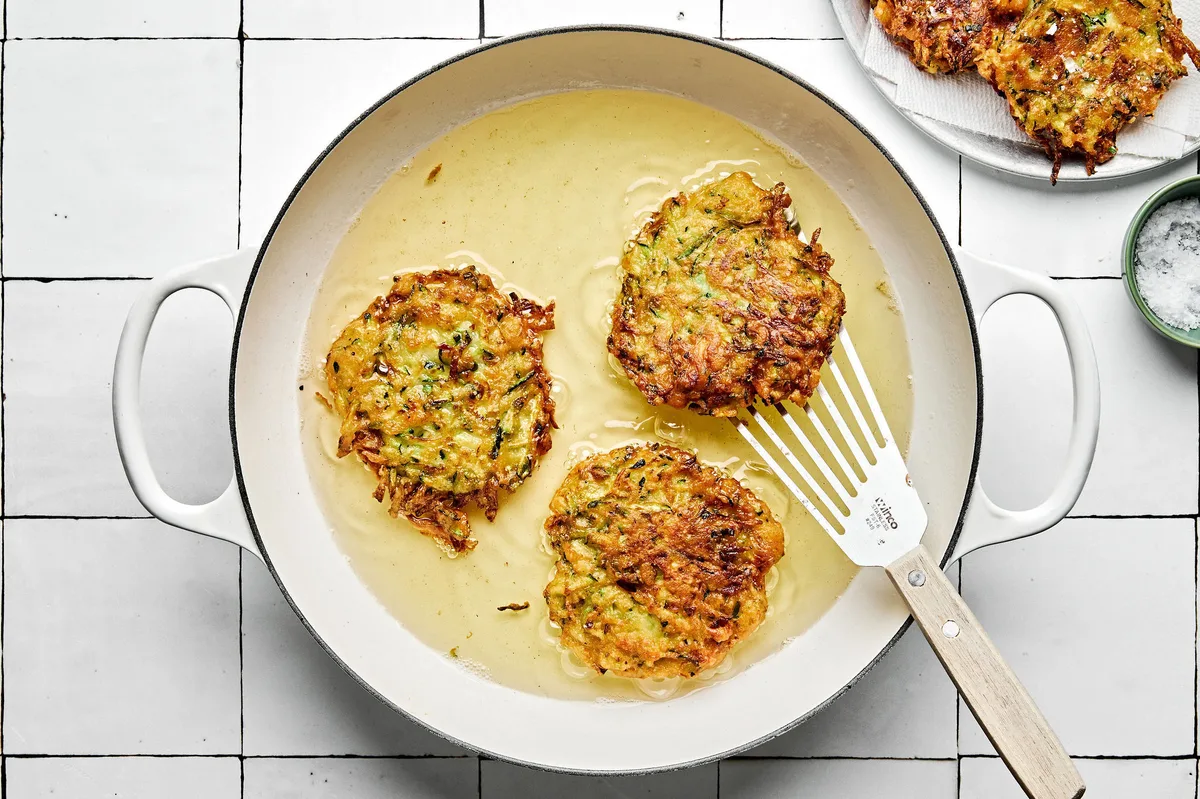 A skillet filled with sizzling oil, actively cooking several zucchini fritters until golden-brown with crispy edges