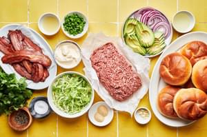 ground beef, brioche buns, avocado, red onion slices, cooked bacon, lettuce, jalapeño and mayonnaise on a yellow surface