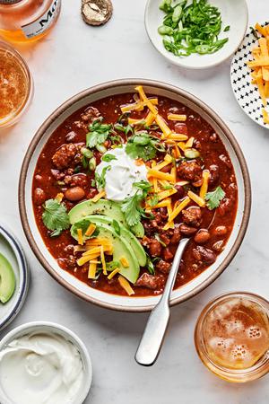 a bowl of homemade chili made with beef, pork, kidney, black and pinto beans topped with cheese, sour cream and avocado
