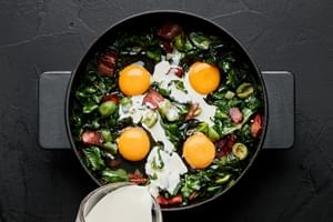 creamed being poured in a skillet with eggs cracked on top of bacon, olives, swiss chard, garlic and onions