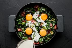 creamed being poured in a skillet with eggs cracked on top of bacon, olives, swiss chard, garlic and onions