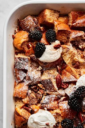 homemade baked french toast made with challah bread in a baking dish topped with dollops of whipped cream and blackberries