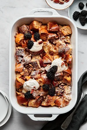 homemade baked french toast made with challah bread in a baking dish topped with dollops of whipped cream and blackberries