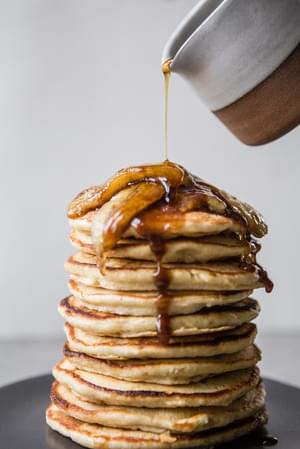 maple syrup being poured over banana pancakes with caramelized bananas and toasted pecans