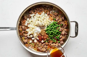 beef stock being added to a skillet with ground beef, onions, carrots, celery, potatoes and spices to make empanada filling