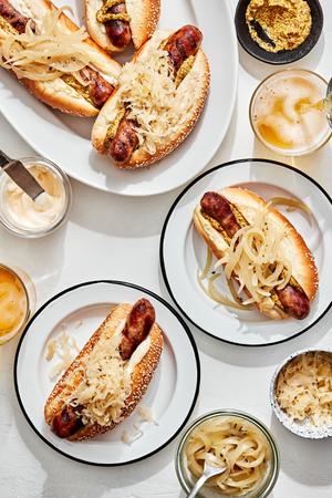 a platter of beer brats served on hoagie rolls topped with sauerkraut, onions, mustard and mayo