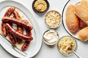 beer brats on a serving plate surrounded by fixings including hoagie rolls, mayo, mustard, onions and sauerkraut