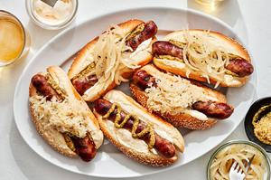 a platter of beer brats served on hoagie rolls topped with sauerkraut, onions, mustard and mayo