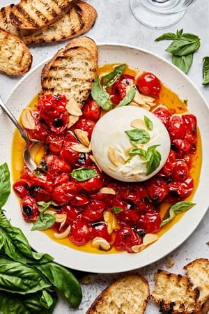 A bowl of braised tomatoes topped with burrata cheese and basil leaves, served with slices of grilled bread on the side