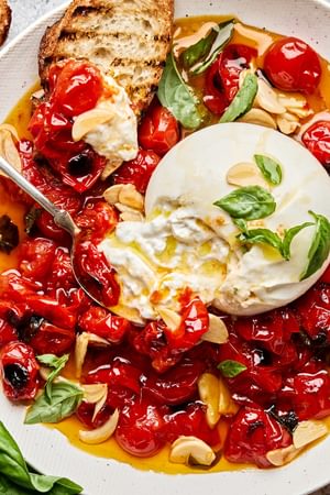 A bowl of braised tomatoes topped with burrata cheese and basil leaves, served with slices of grilled bread on the side