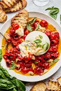 A bowl of braised tomatoes topped with burrata cheese and basil leaves, served with slices of grilled bread on the side