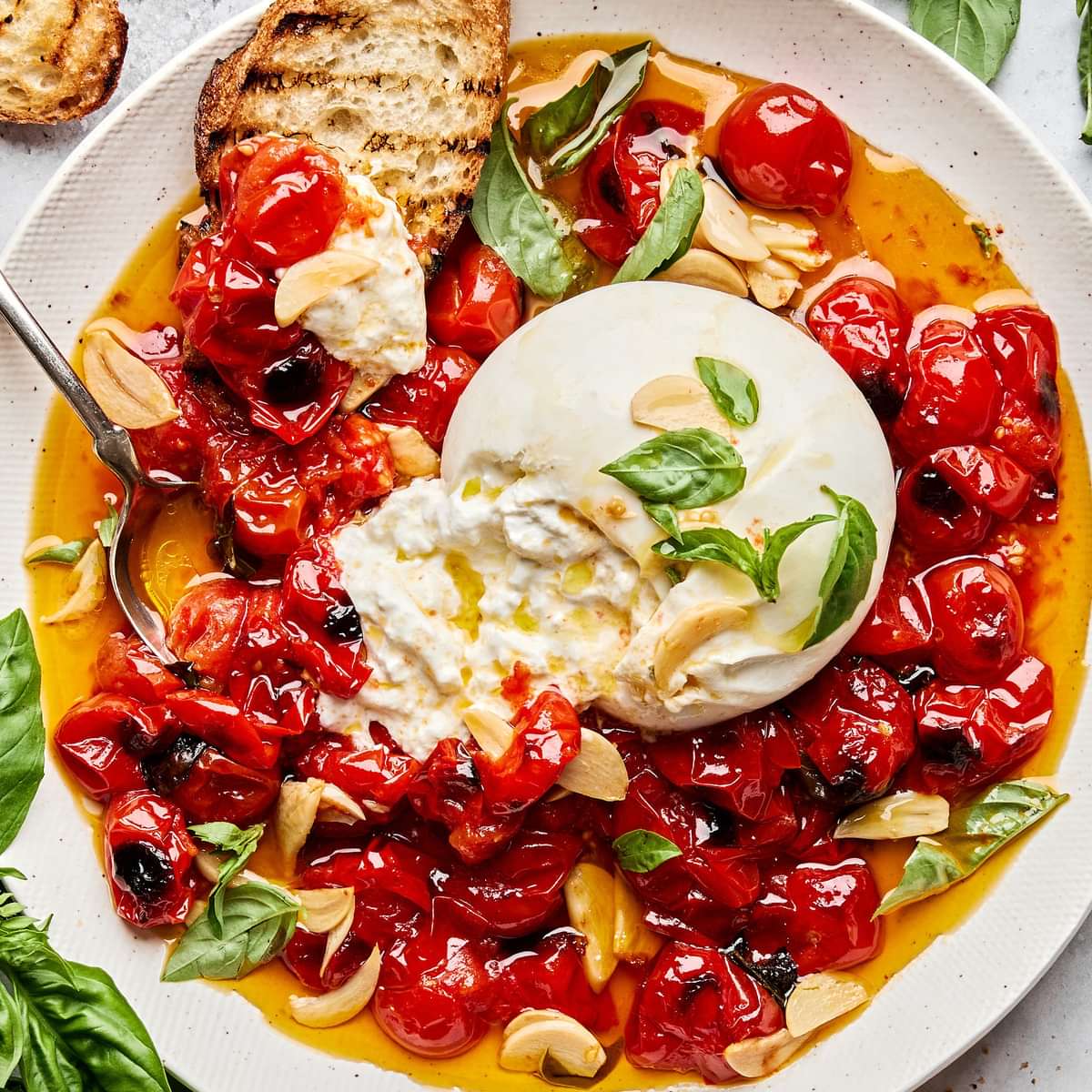 A bowl of braised tomatoes topped with burrata cheese and basil leaves, served with slices of grilled bread on the side