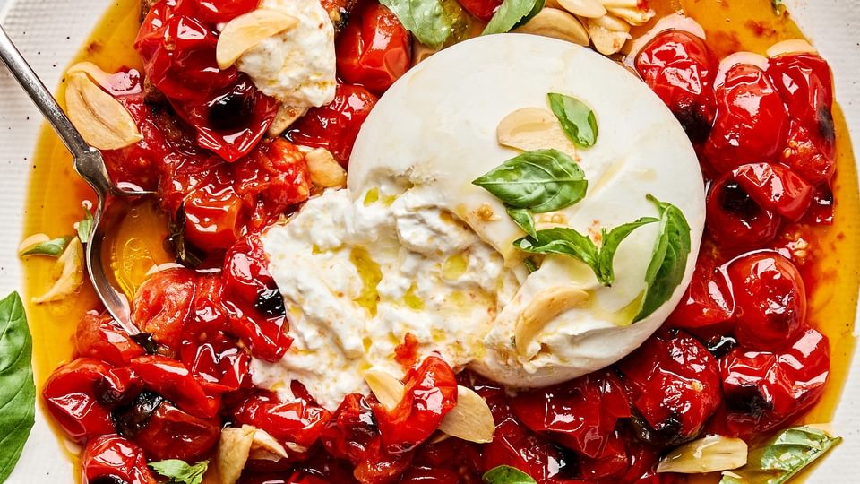A bowl of braised tomatoes topped with burrata cheese and basil leaves, served with slices of grilled bread on the side