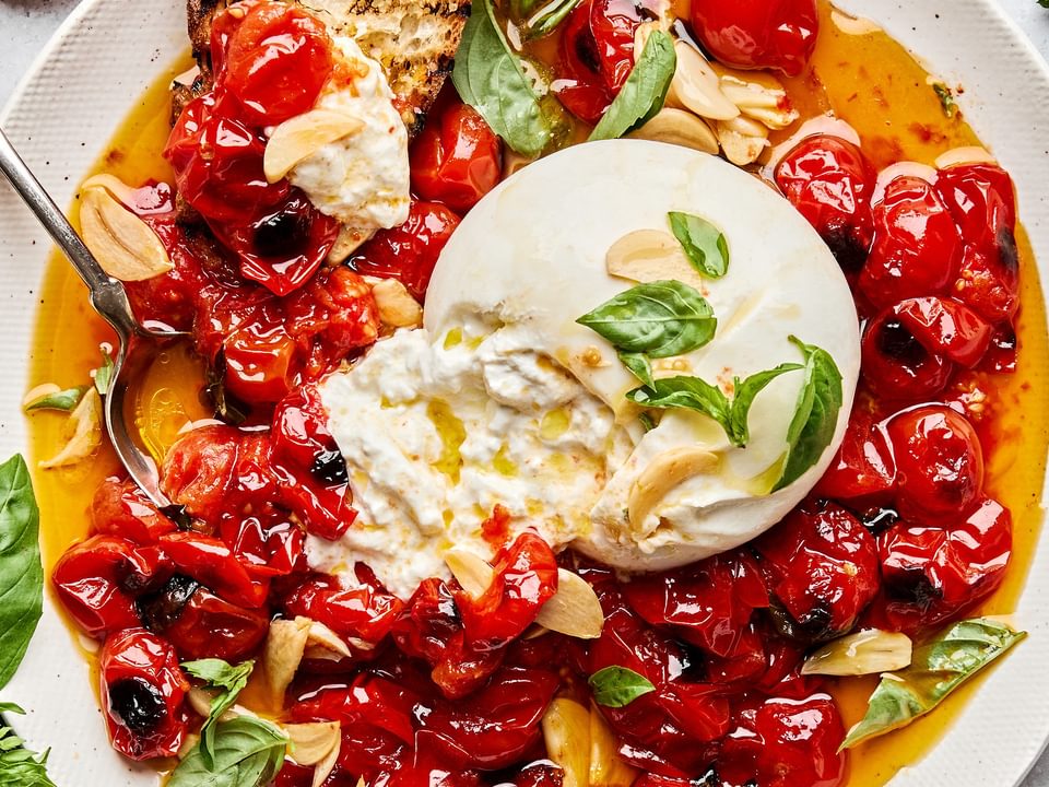 A bowl of braised tomatoes topped with burrata cheese and basil leaves, served with slices of grilled bread on the side