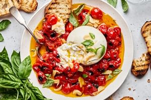 A bowl of braised tomatoes topped with burrata cheese and basil leaves, served with slices of grilled bread on the side