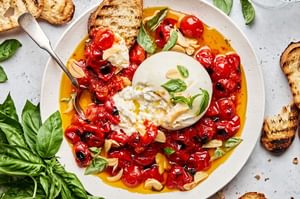 A bowl of braised tomatoes topped with burrata cheese and basil leaves, served with slices of grilled bread on the side