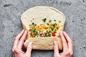 a breakfast burrito being rolled filled with sausage, eggs, potatoes, cilantro, avocado and salsa
