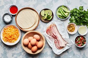 eggs, bacon, cheese, chiles, salt, corn tortillas, cilantro, avocado, pico de gallo, limes and hot sauce in prep bowls