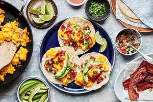 breakfast tacos being assembled surrounded by all the fixings: eggs, bacon, salsa, avocado, cilantro, limes and hot sauce
