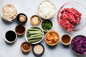 raw thinly sliced steak, onions, kimchi, red cabbage, cucumbers, bean sprouts and spices in prep bowls for beef bulgogi