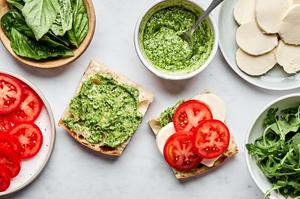 a caprese sandwich being assembled with creamy pesto, mozzarella, tomato, arugula, pesto and balsamic glaze