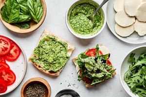 a caprese sandwich being assembled with creamy pesto, mozzarella, tomato, arugula, pesto and balsamic glaze