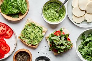 a caprese sandwich being assembled with creamy pesto, mozzarella, tomato, arugula, pesto and balsamic glaze