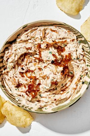 a bowl of caramelized onion dip made with cream cheese, sour cream, spices and chives surrounded by potato chips