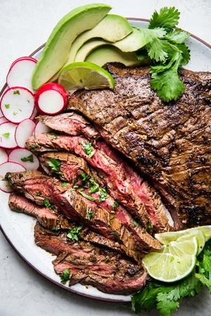 A plate of grilled carne asada with tortillas, limes, avocado and radishes