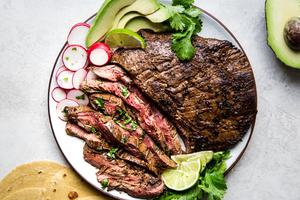 A plate of grilled carne asada with tortillas, limes, avocado and radishes