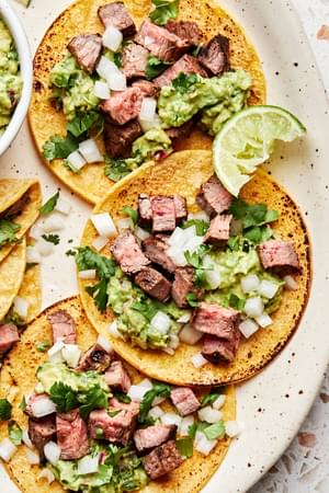 a plate of carne asada tacos topped with cilantro, onion, and guacamole served with a lime wedge