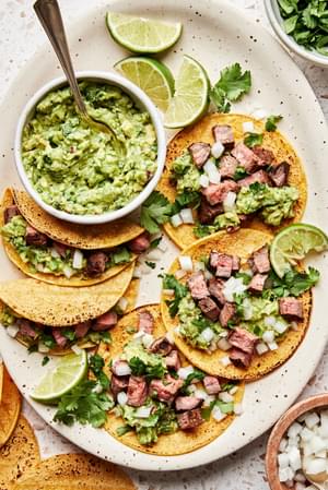 a plate of carne asada tacos topped with cilantro, onion, and guacamole served with a lime wedge