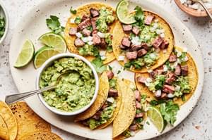 a plate of carne asada tacos topped with cilantro, onion, and guacamole served with a lime wedge