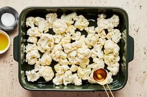 raw cauliflower florets being tossed with olive oil and salt in a baking dish