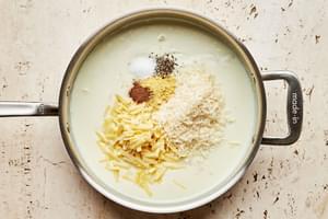 mustard powder, salt, pepper, nutmeg, Parmesan, and Gruyère being added to a roux in a skillet